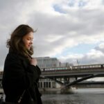 A young woman in a winter coat smiling while walking by a bridge in Berlin.